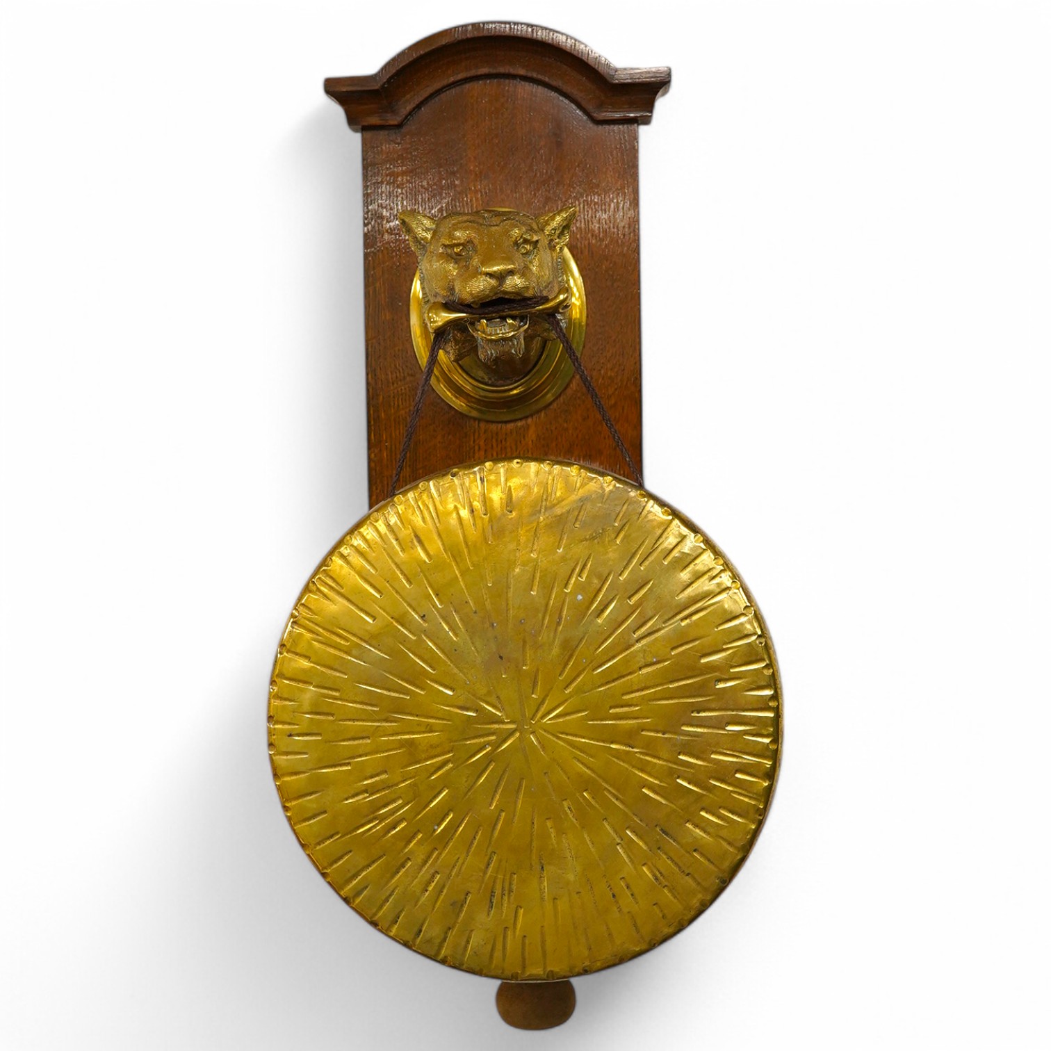 An early 20th century brass wall hanging gong, the holder in the form of a big cat, mounted on an oak plinth, the gong is supported on a bone clenched between its teeth, with the beater on a separate hook behind, 55cm to